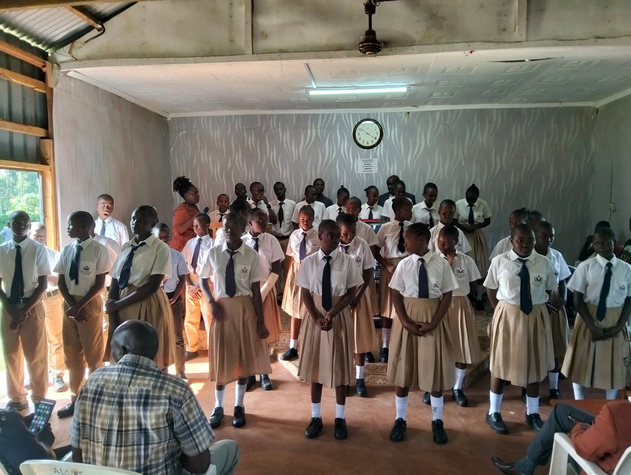 Students in white shirts and tan uniforms standing in rows inside a room for assembly.