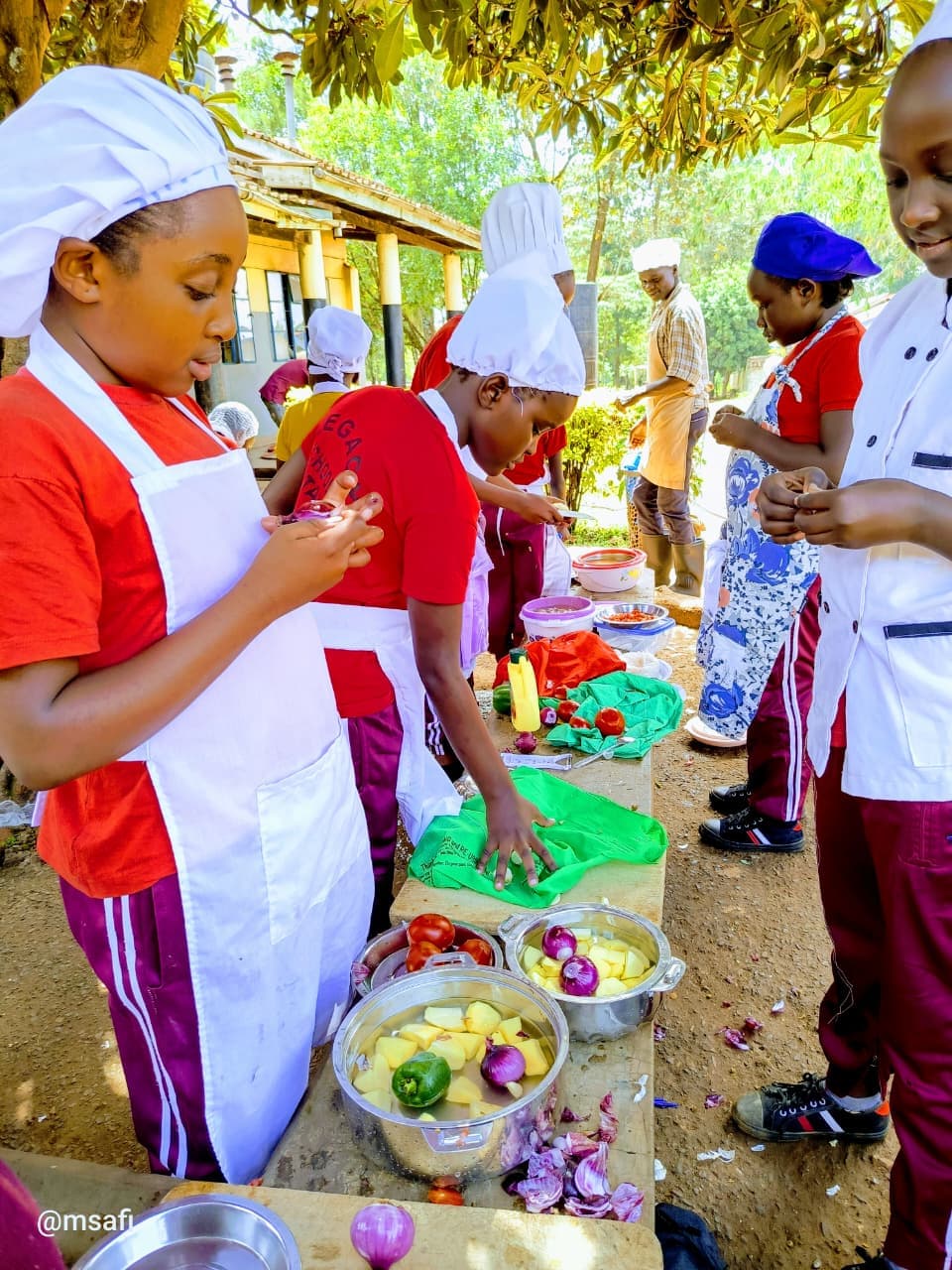 Students in chef hats and aprons prepare fresh vegetables at an outdoor cooking station.