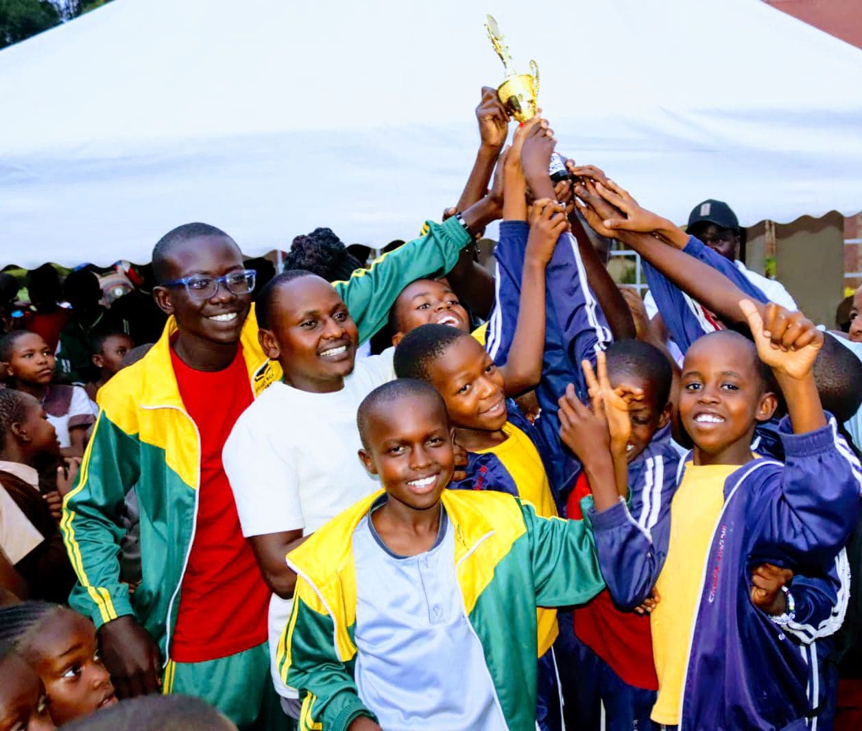 Smiling children and adults in tracksuits celebrate a victory by holding up a gold trophy.