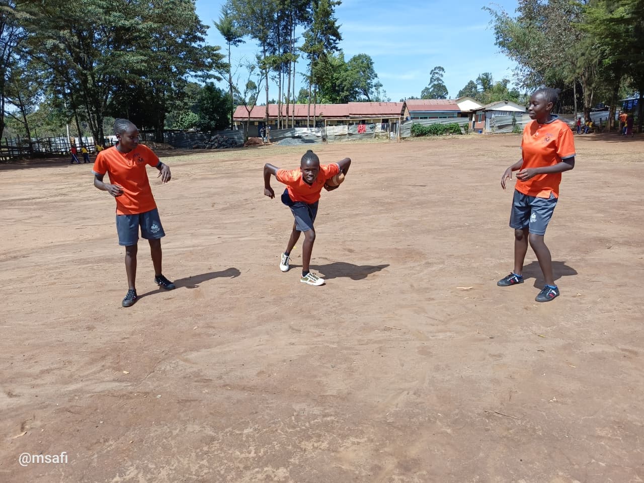A girl runs with a rugby ball while two teammates watch on a dusty field.