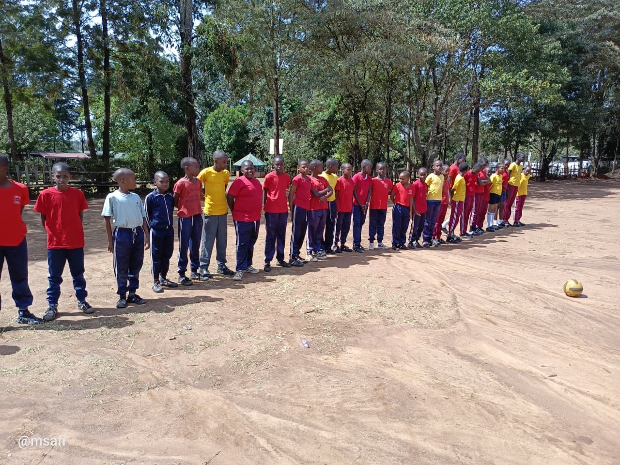 Boys in red and yellow shirts line up on a dirt field with soccer ball.