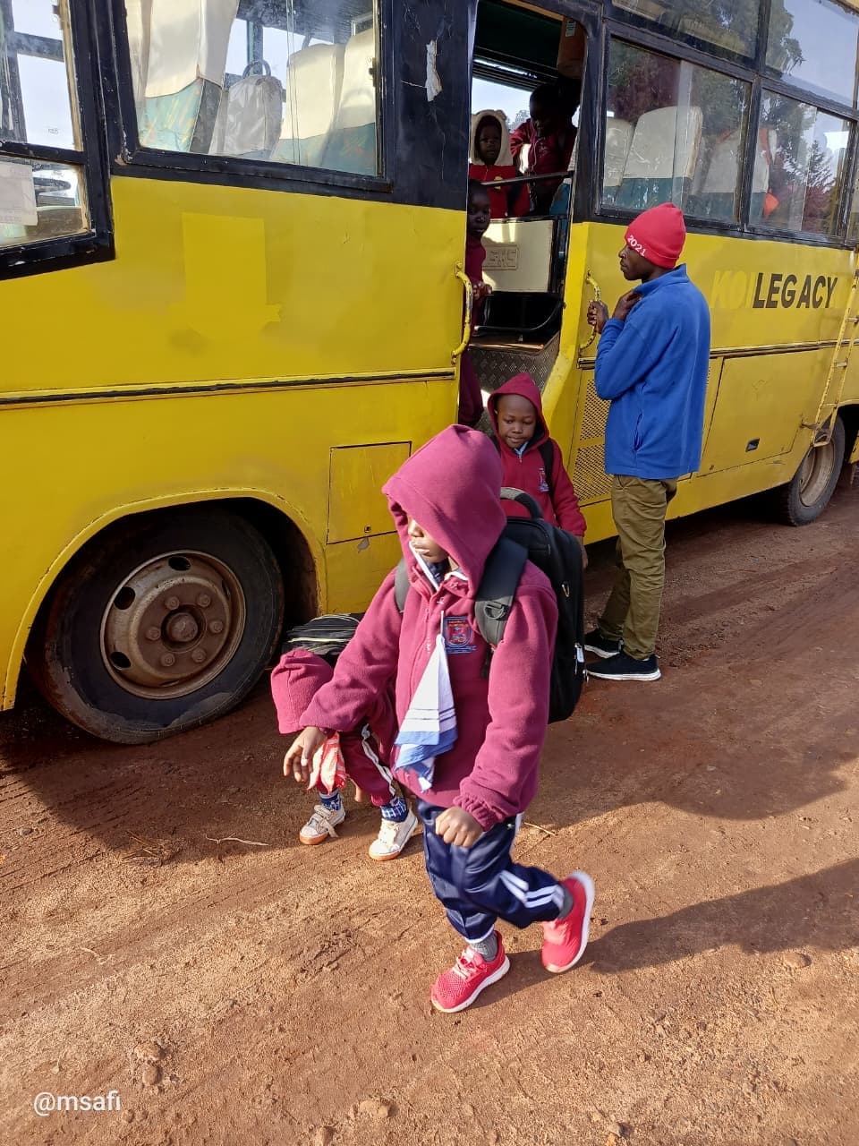 Children in maroon school uniforms disembarking from a yellow bus on a dirt road.