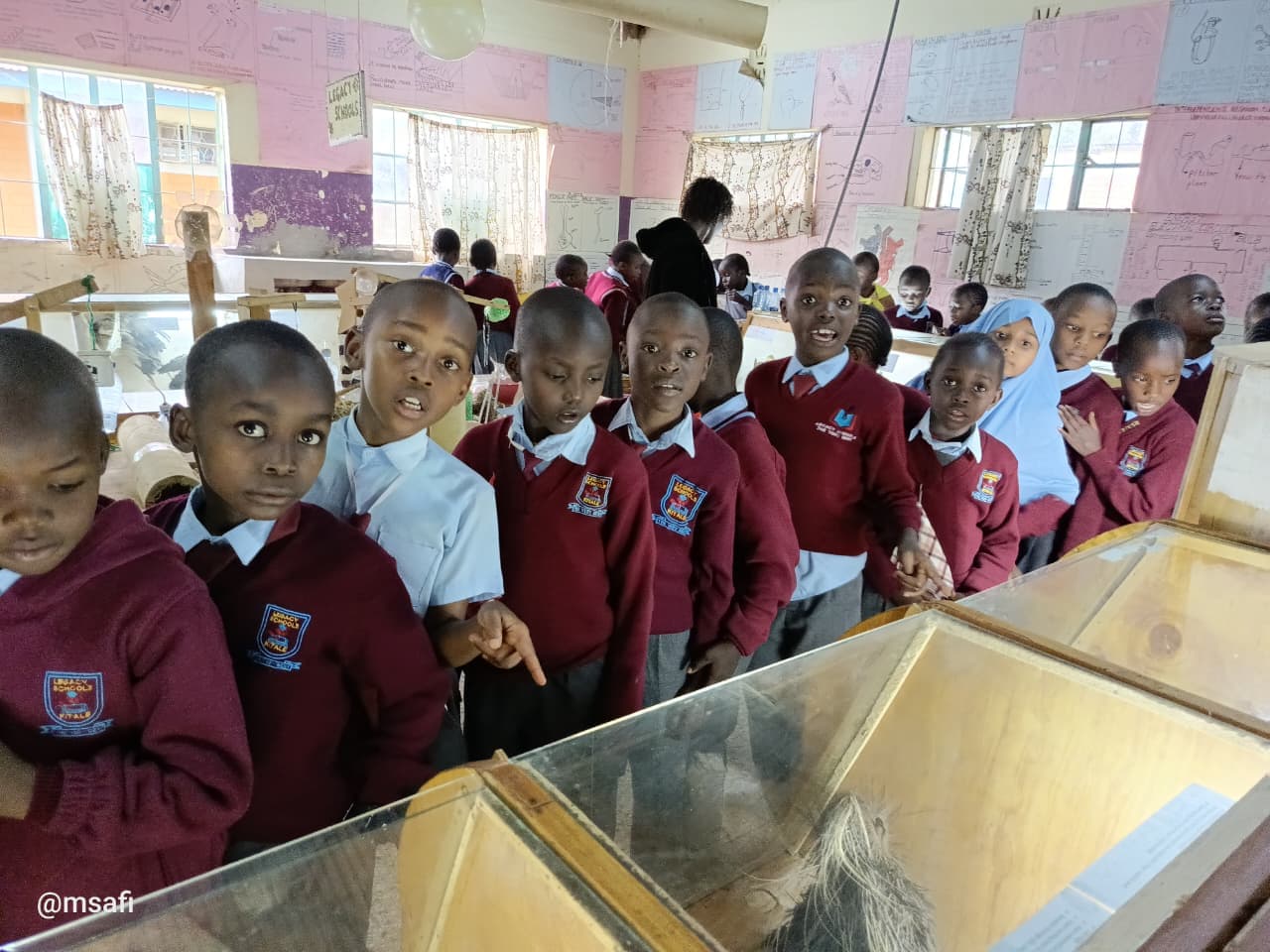 Schoolchildren in maroon uniforms look curiously at educational displays inside a brightly lit classroom.