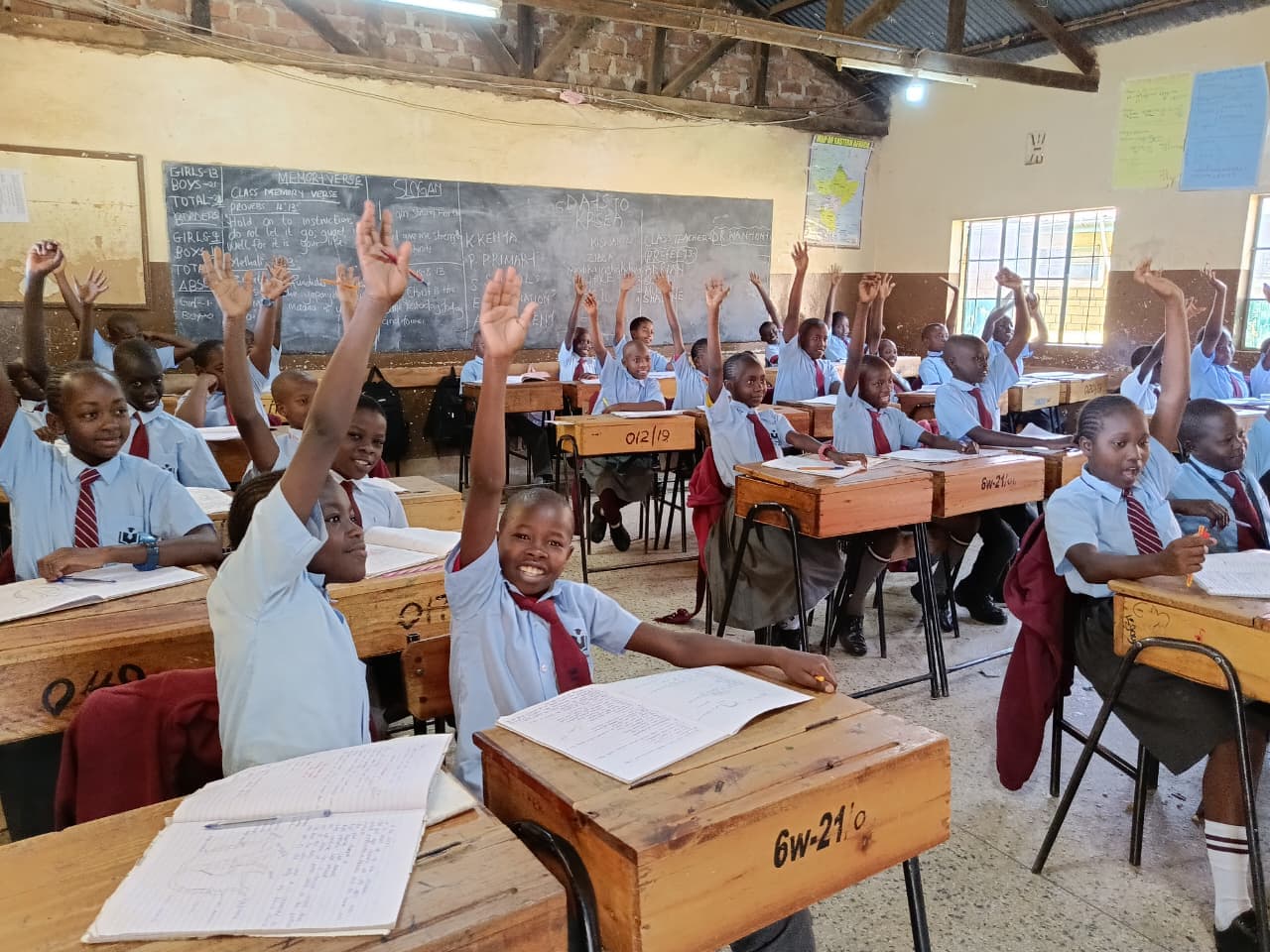 Students in blue uniforms and red ties raise their hands at wooden school desks.
