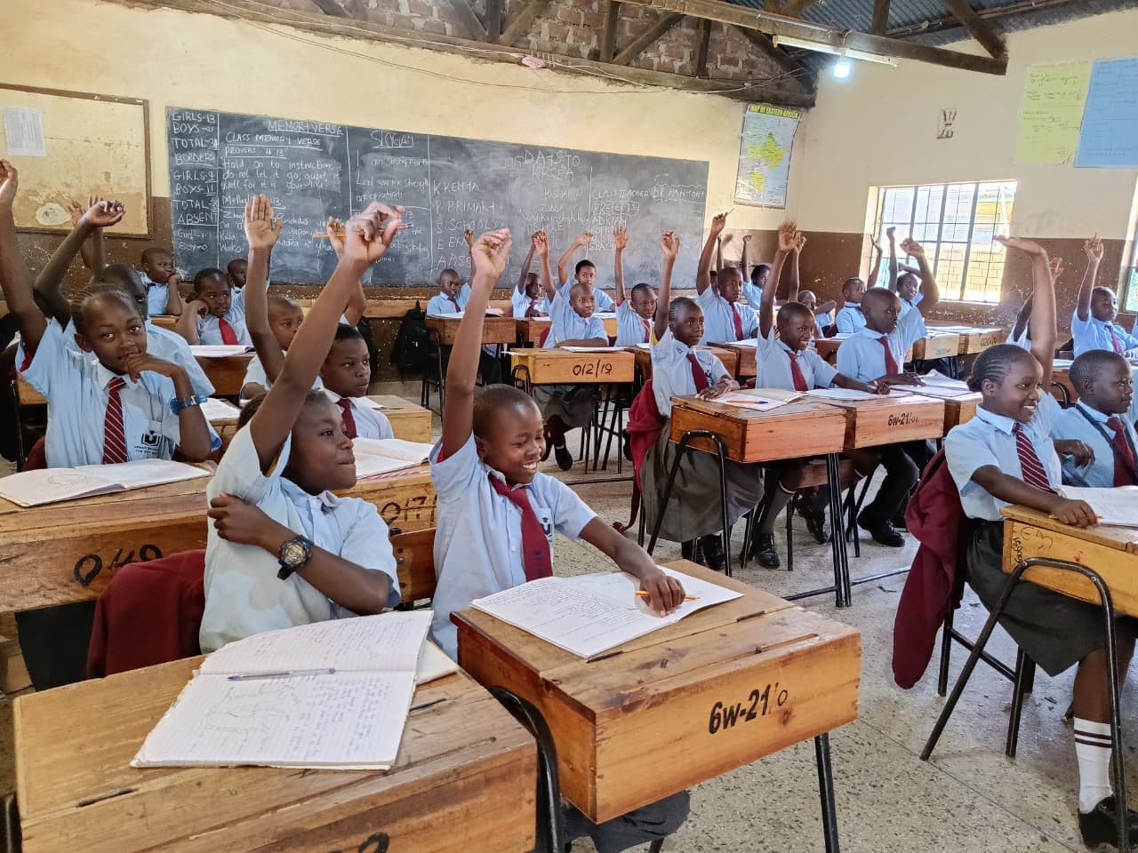 Students in blue uniforms eagerly raise their hands at wooden desks in a classroom.