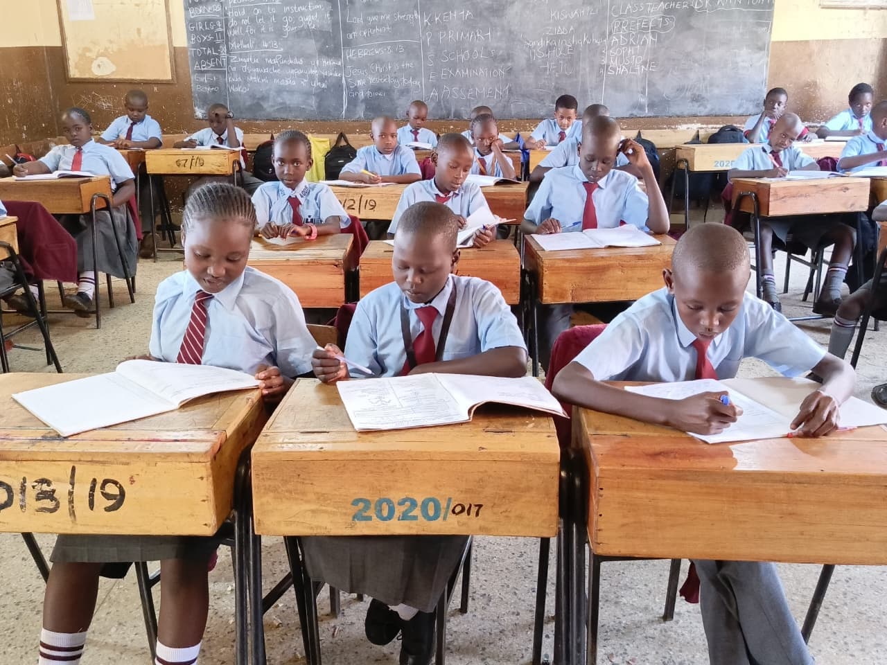 Students in blue uniforms and red ties sit at wooden desks, writing in a classroom.
