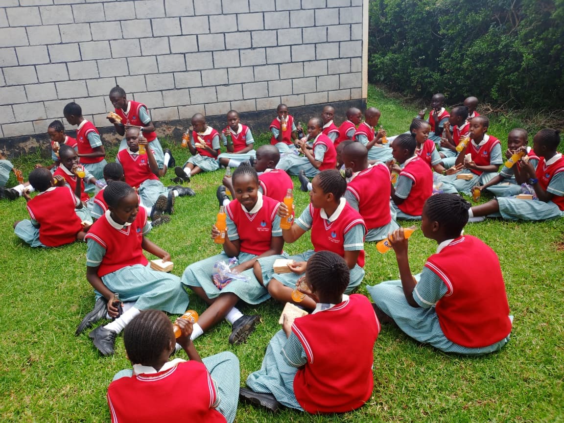 Schoolgirls in red vests sit on grass, eating sandwiches and drinking orange soda together.