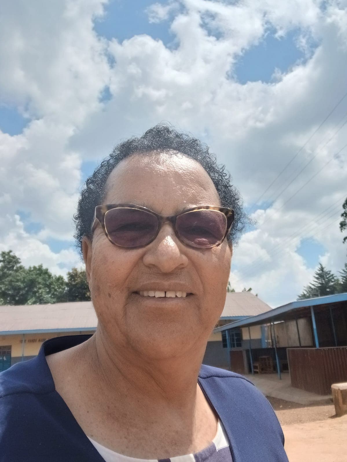 Smiling woman with short curly hair and sunglasses standing outdoors under a bright, cloudy sky.
