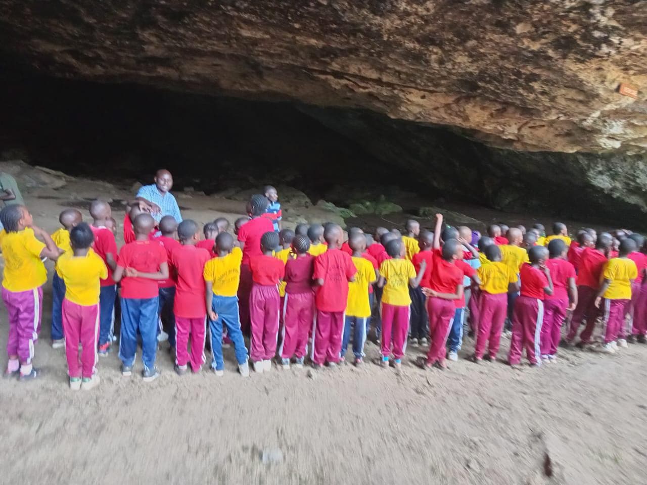 School children in red and yellow uniforms listen to a guide inside a large cave.